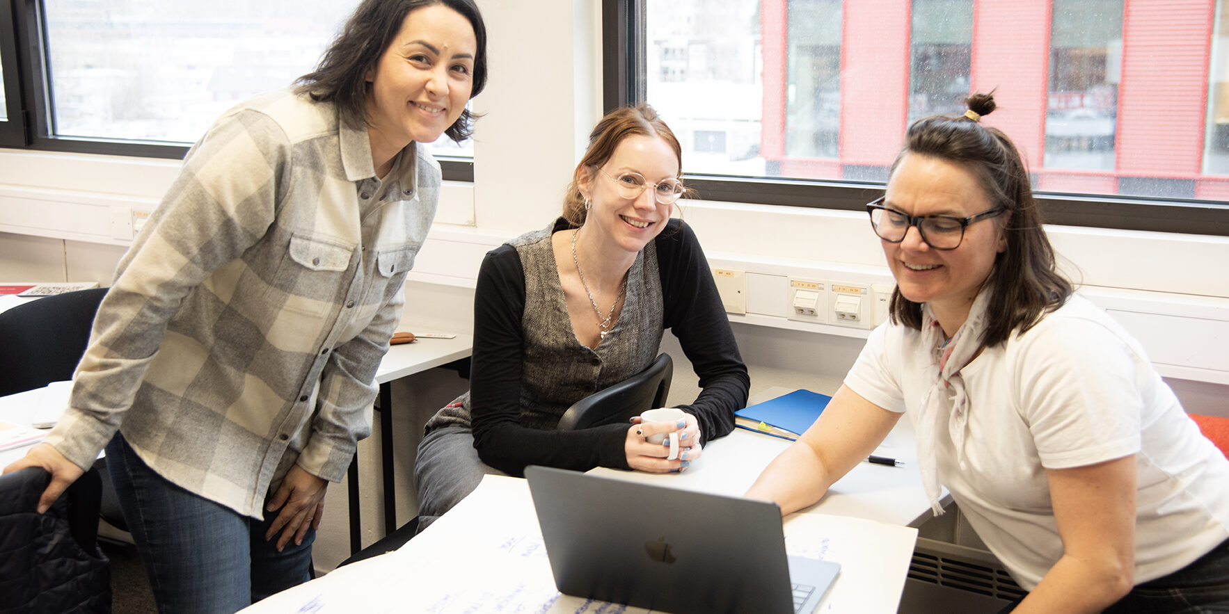 Tre smilende damer sitter rundt et bord og kikker på en laptop